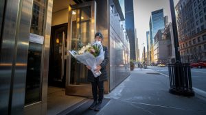 Manhattan street scene early morning, female florist delivery person with wrapped sympathy flowers at building entrance, professional urban photography, towering skyscrapers background, dramatic morning light, clear day